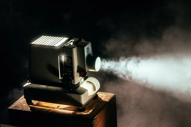 A film projector  sits on a small table, lit up and projecting light. There's also hazy smoke in the room. 