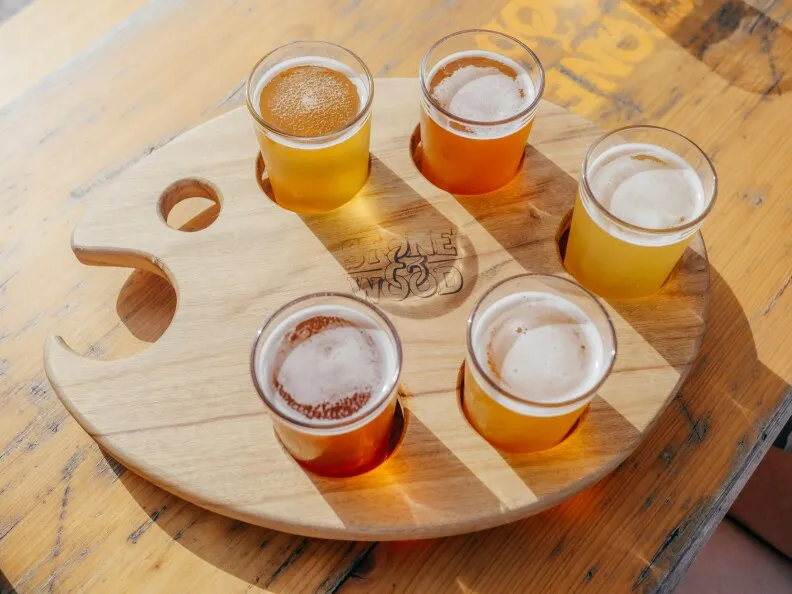 image of five different types of beers. Looking door at the beers sitting on top of a wooden table. The beers are also in a unique beer holding tray that's shape like a painting tray.