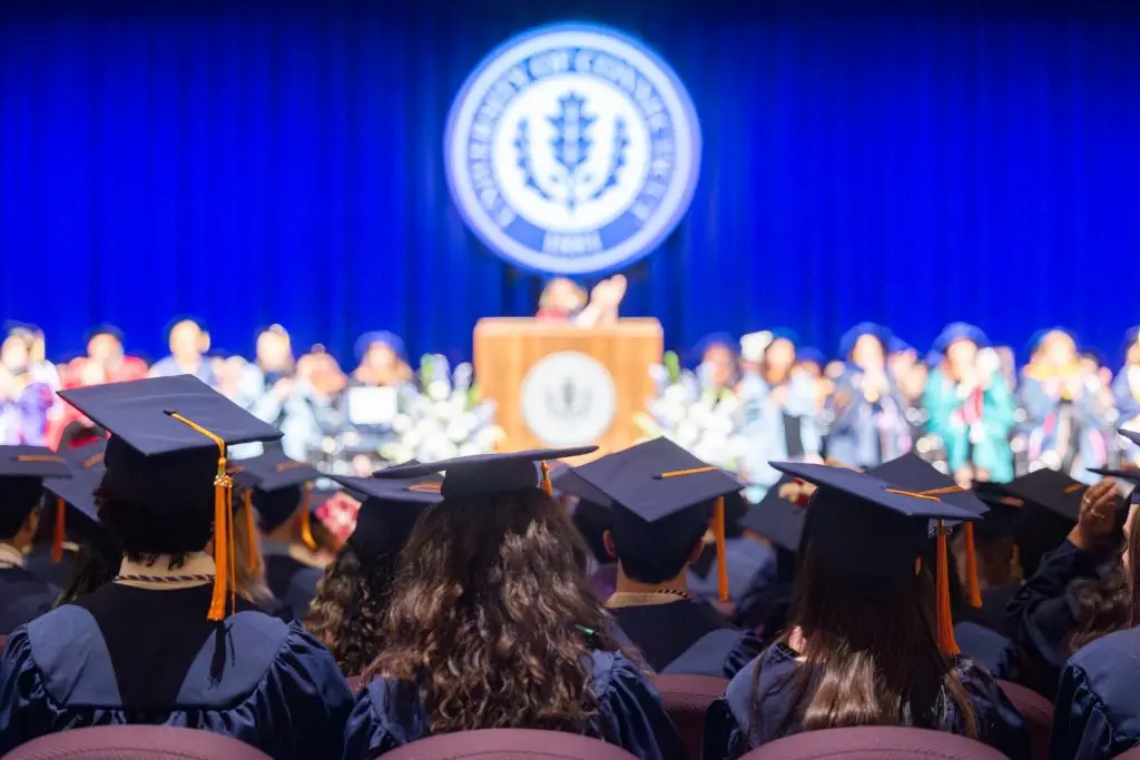 Graduates in their caps and gowns listening to their commencement speaker with delgates on stage and a speaker at the mic