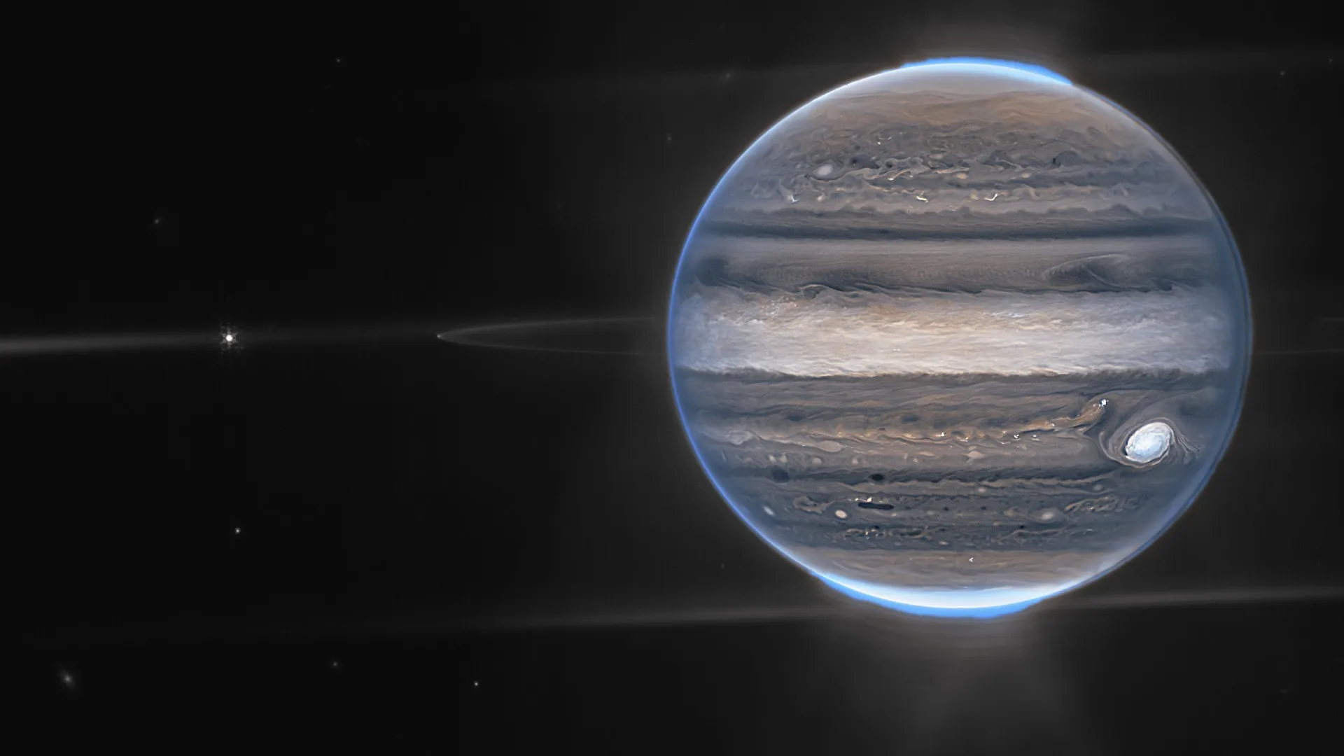 Jupiter looks like a glowing navy blue marble in space with stripes of glowing beige, white and black. A large spot on it is glowing pale blue and its poles indicate bright auroras.