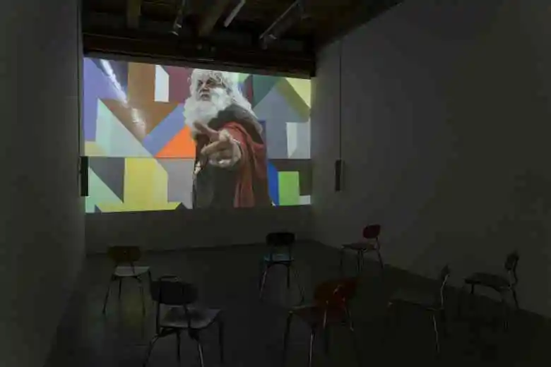 In a darkened viewing room, with chairs set up before a screen, is a projected image of the artist in old fashioned garb, wearing a large white beard and long white wig. He points angrily toward the viewer. Behind him is a colorful geometric pattern on the wall.