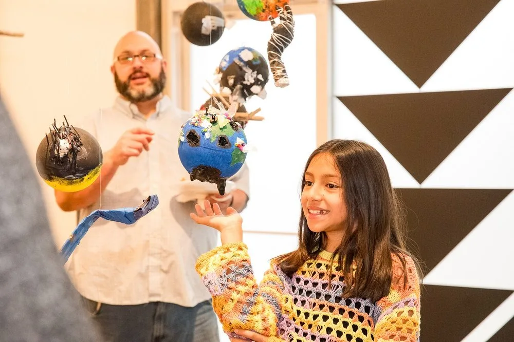 A middle school student poses with her artwork, a mobile with planet-esque shapes hanging from the ceiling.