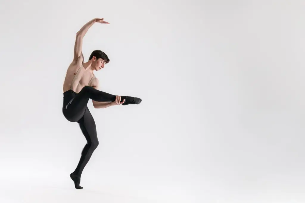 A male dancer poses, right arm arched overhead, right leg grasped, at the ankle, by left hand.