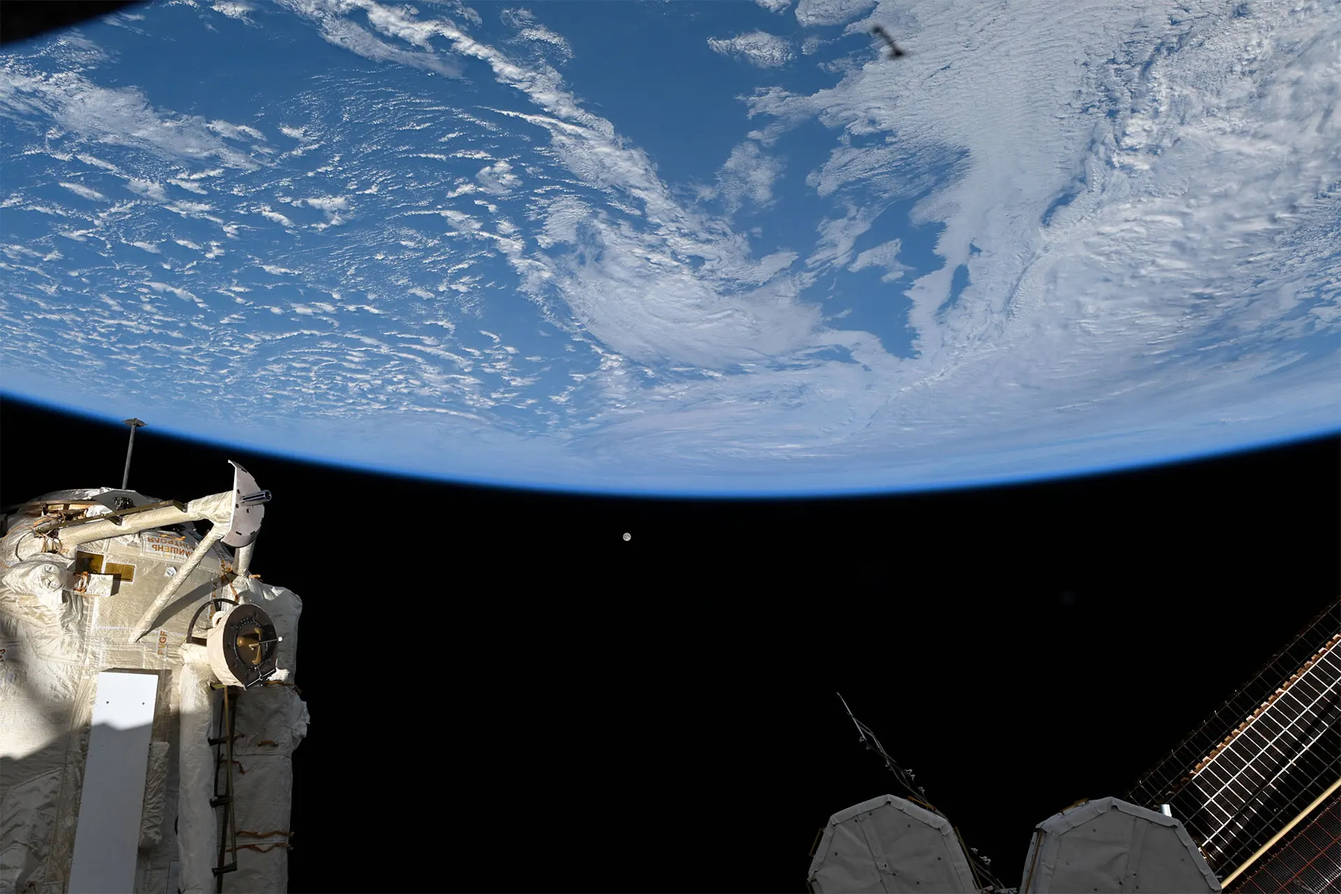 a full moon looks tiny set against the backdrop of the blackness of space with Earth's horizon and a space station in the foreground..