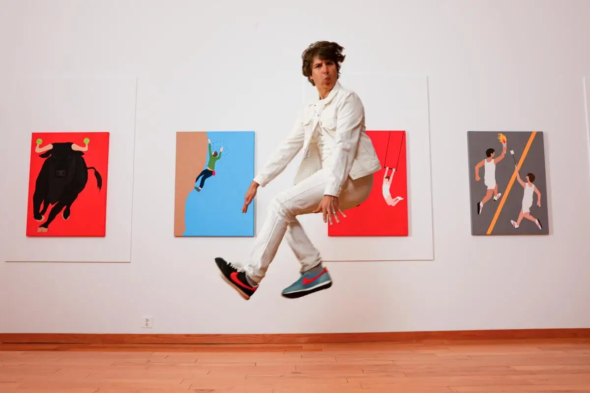 Demetri Martin, in a white suit, jumps in front of a white gallery wall with four colorful paintings. 