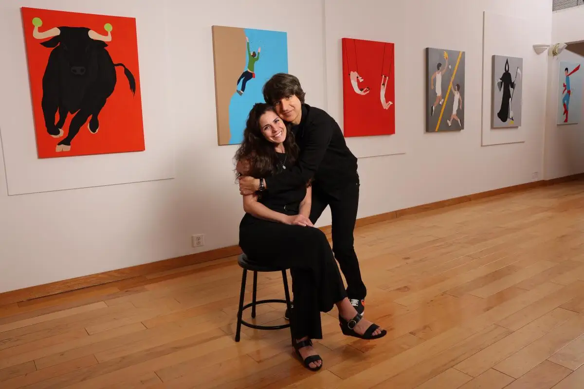 Demetri Martin hugs his wife, Rachael Beame Martin, seated on a stool in front of colorful paintings.