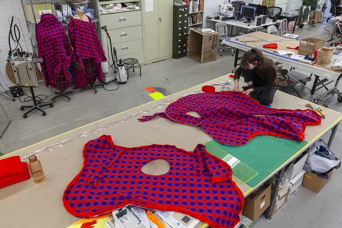 Movement towards Freedom - Brendan Fernandes at the Fabric Workshop and Museum 3 A birds-eye view of a workshop in a museum shows a woman in black with long brown hair sitting on a tabletop sewing a border on a bright colored, red- and-blue checkered costume to be worn in a performance.