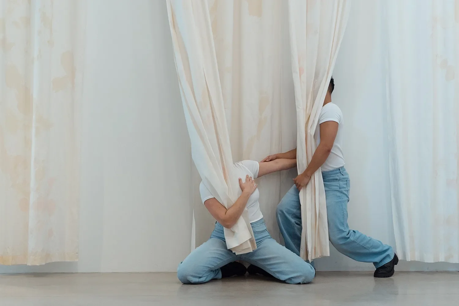 Movement towards Freedom - Brendan Fernandes at the Fabric Workshop and Museum 4 Two performers in light-colored jeans and white t-shirts perform a dance movement together, while partly hiding behind beige and white curtains.