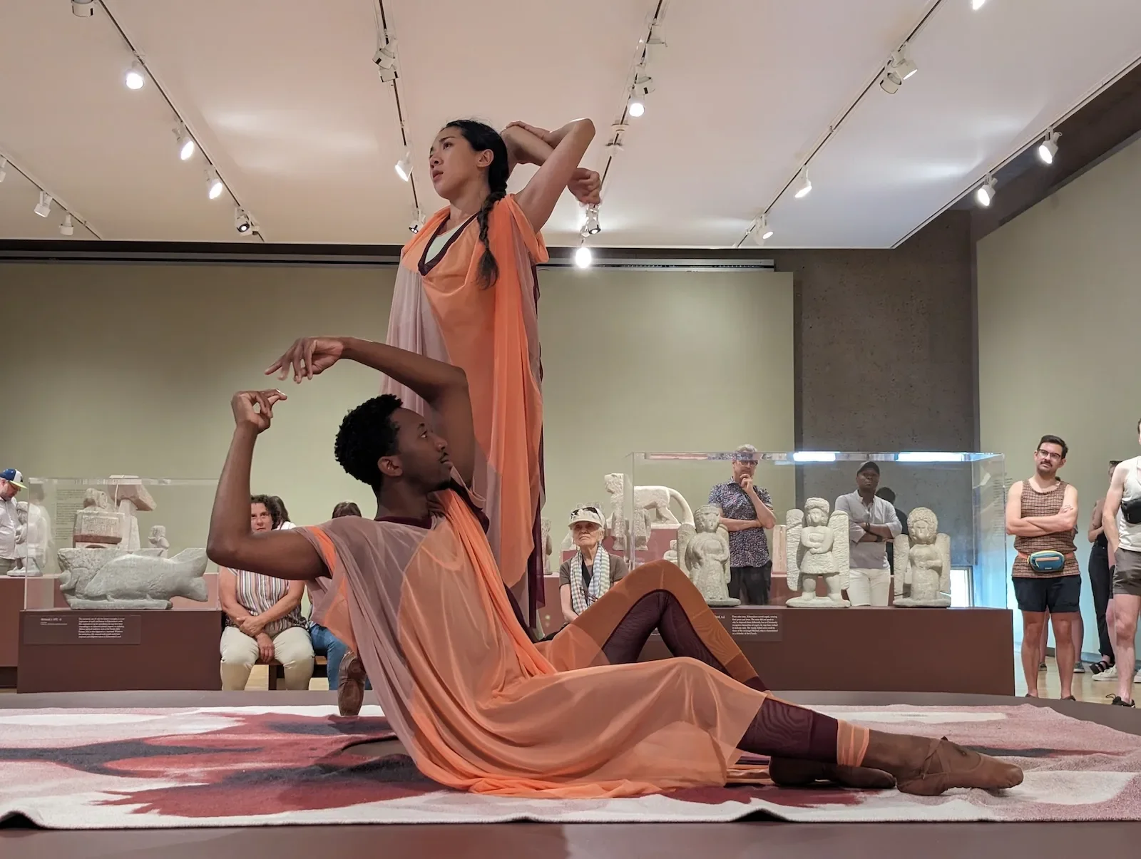 Movement towards Freedom - Brendan Fernandes at the Fabric Workshop and Museum 5 Two performers, a man, on the floor, and a woman standing next to him wear peach and brown, Greek-like gowns and approximate statues in their pose, the man with arms above and encircling his head and the woman’s arms crossed above and in back of her head. An audience looks on.