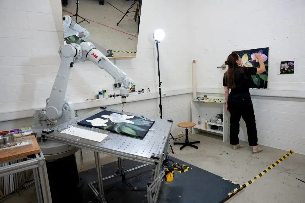 view of an artist painting in the studio and a robotic arm painting on a canvas on a table nearby