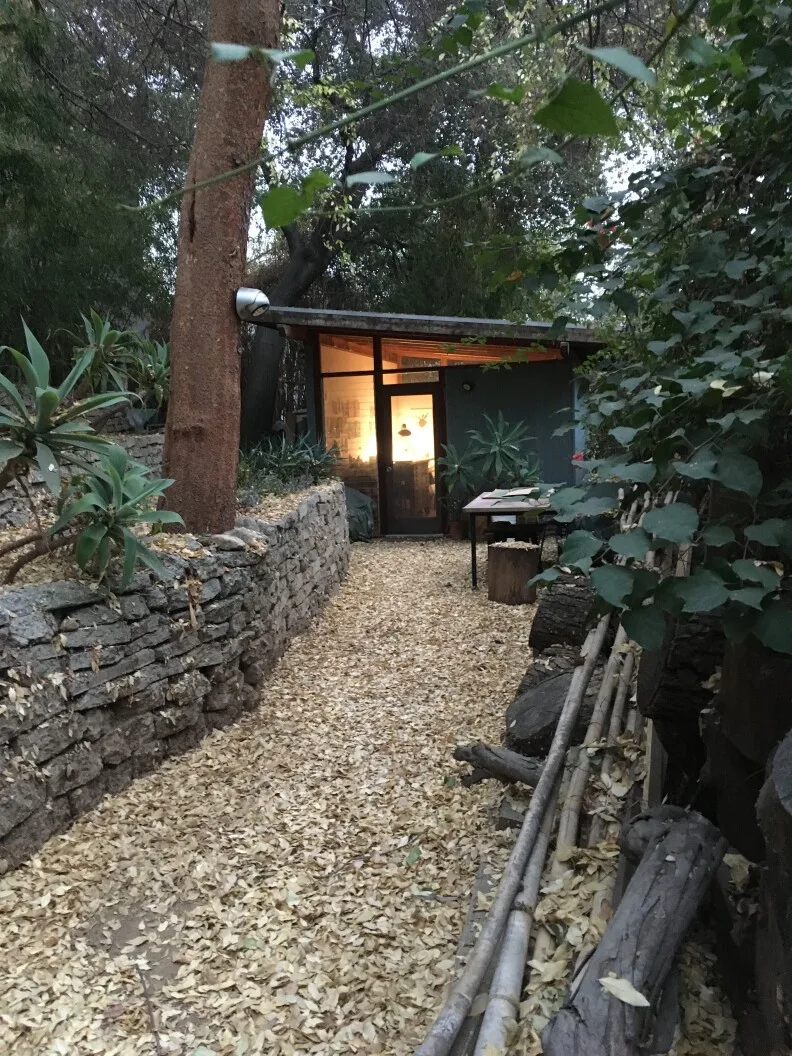 A one story, one room building, with a light shining through the window, surrounded by trees.