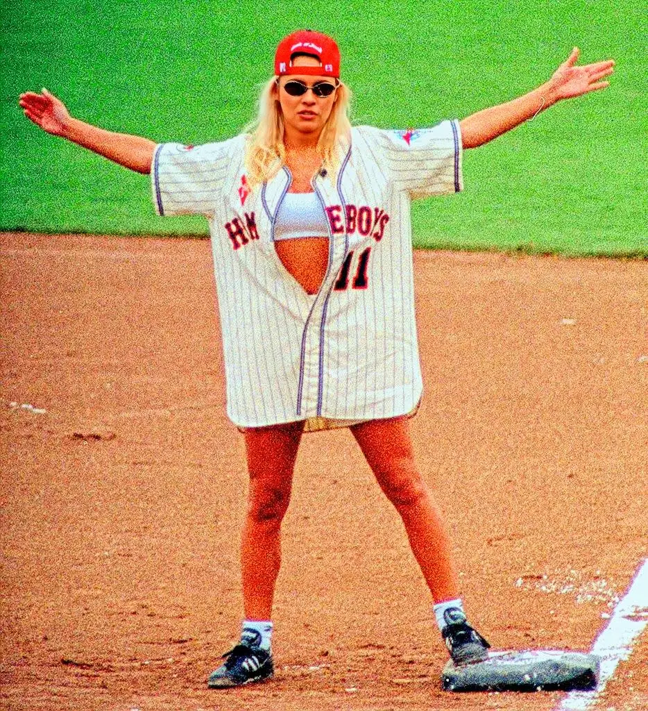 Person on a baseball field wearing a loose-fitting jersey, shorts, and cap, gesturing with arms wide open