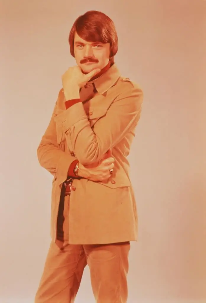 Person posing with hand on chin, wearing a safari-style jacket, long sleeves, and pants, with a thoughtful expression