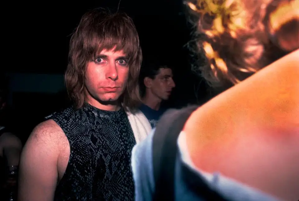A person with a surprised expression, wearing a sleeveless patterned top, stands indoors at an event
