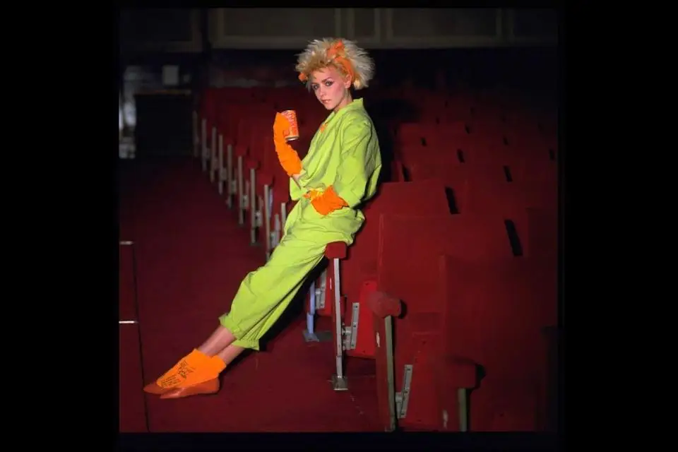 Person in avant-garde outfit sitting sideways on theater seat, wearing vibrant, eclectic clothing and striking a pose