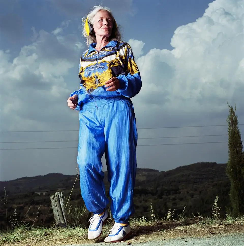 Older person outdoors, wearing a stylish, patterned tracksuit and sneakers, appearing joyful and energetic. Background shows a hilly landscape and cloudy sky