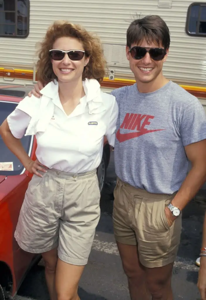 Two individuals stand smiling outdoors, both in casual attire with sunglasses. One wears a branded shirt, the other has a towel over their shoulder