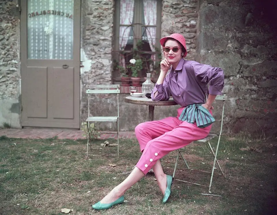 Person in stylish vintage outfit, seated outdoors, wearing a wide-brim hat, blouse with ruffled waist, cropped pants, and flats