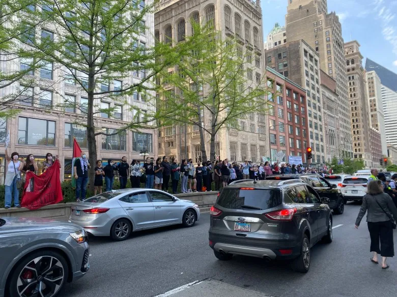 A long row of protesters stand on the median of Michigan Ave in downtown Chicago, protesting the Art Institute of Chicago and SAIC for their complicity in the genocide of Gaza. Tall buildings can be seen behind them, and traffic flows north in several lanes in front of them.