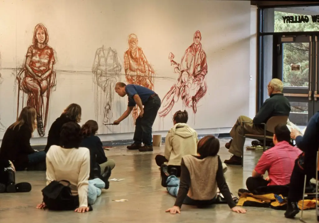 artists drawing seated figure in a chair while students watch on from a cement floor