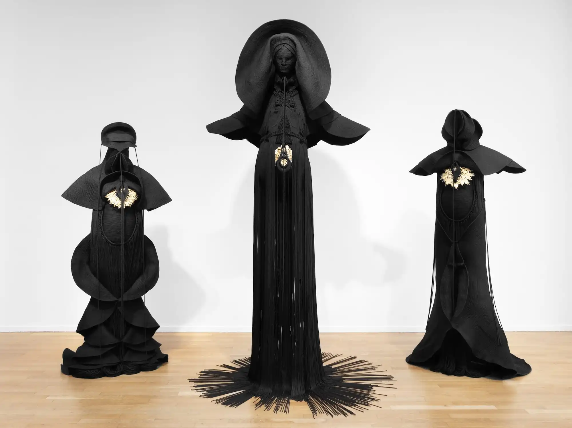 Installation view of three sculptures made from black cord, thread, and metal in the form of figures or long, dramatic garments