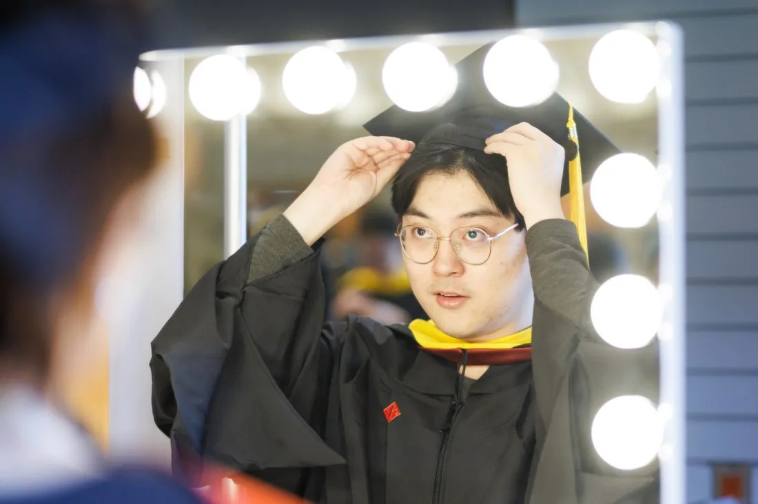 A graduate in cap and gown adjusts their mortarboard while looking in a Hollywood-style vanity mirror with bright bulbs around the frame.