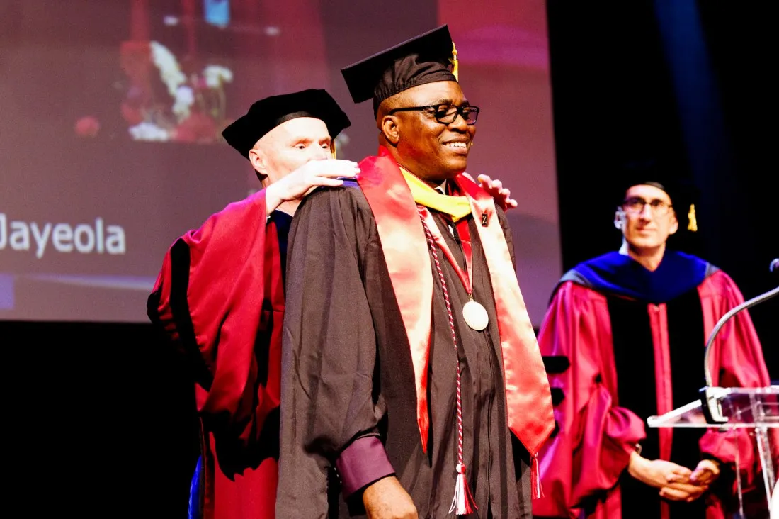 Faculty in colorful academic regalia (red, purple, and blue robes) stand together on stage, with one person adjusting the regalia and medal that a graduate is wearing around their neck.