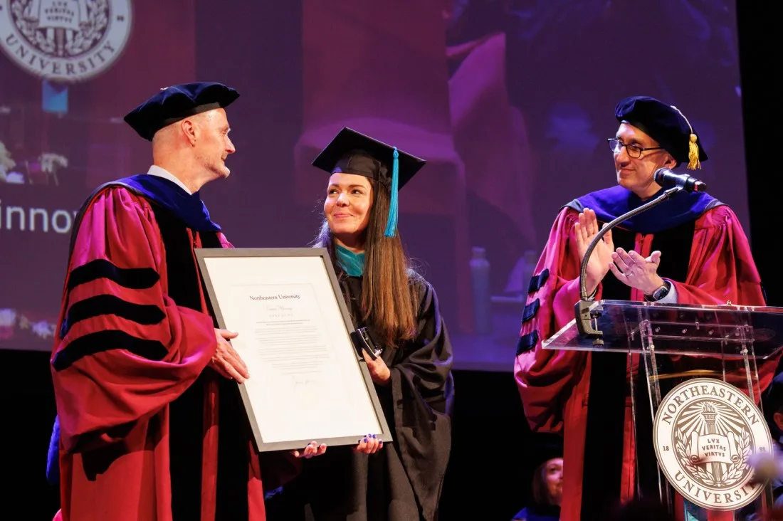 A dean in red and black academic robes presents a framed certificate to a female graduate in a black cap and gown, while another official in purple robes applauds nearby.
