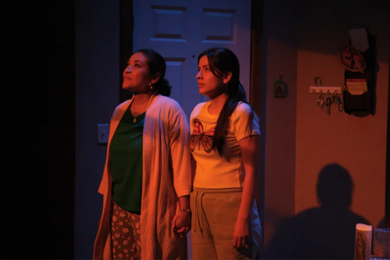 Two women on a stage,in darkness, with long, dark hair.