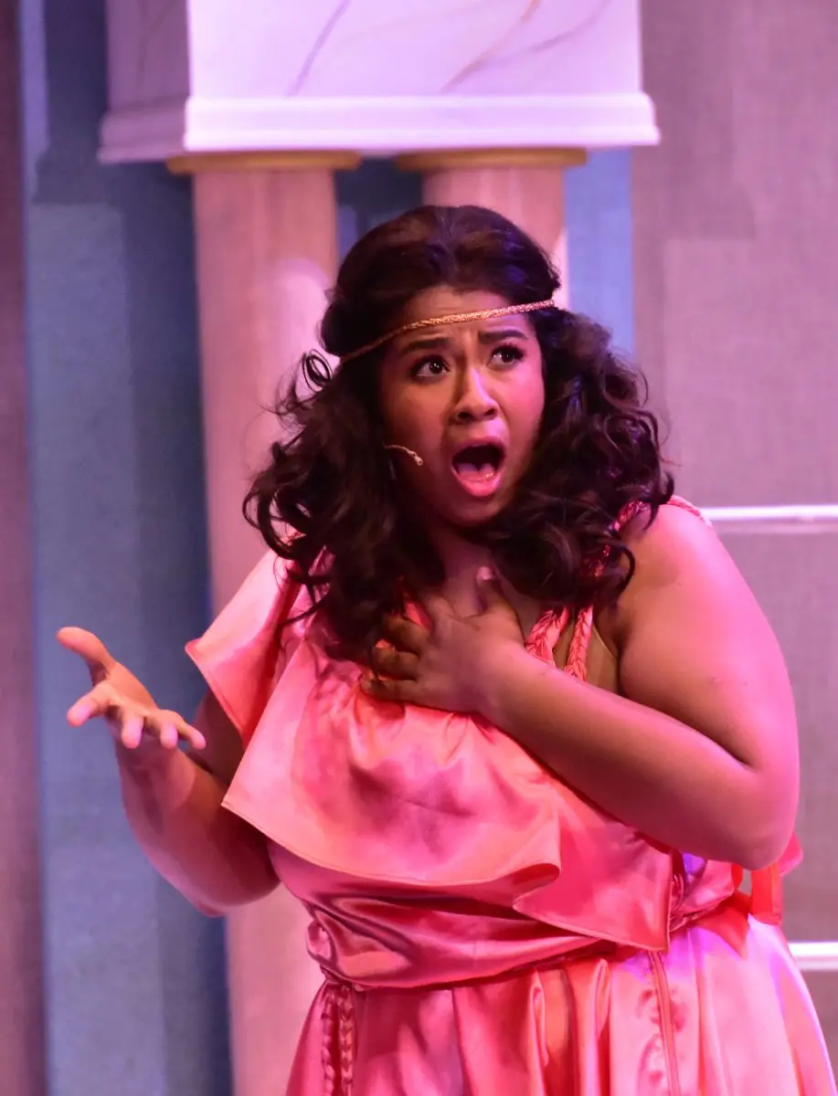 A woman in a pink costume with a headband stands on stage, looking surprised with her mouth open and one hand on her chest and the other raised.