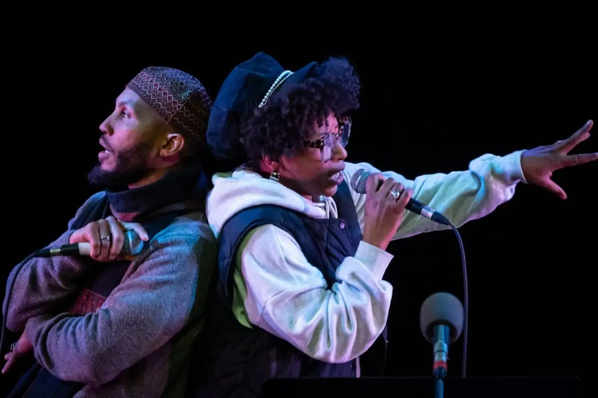 Two people stand back-to-back on stage, holding microphones and performing. One wears a hat and scarf, the other has sunglasses and a cap, pointing outward with one hand.