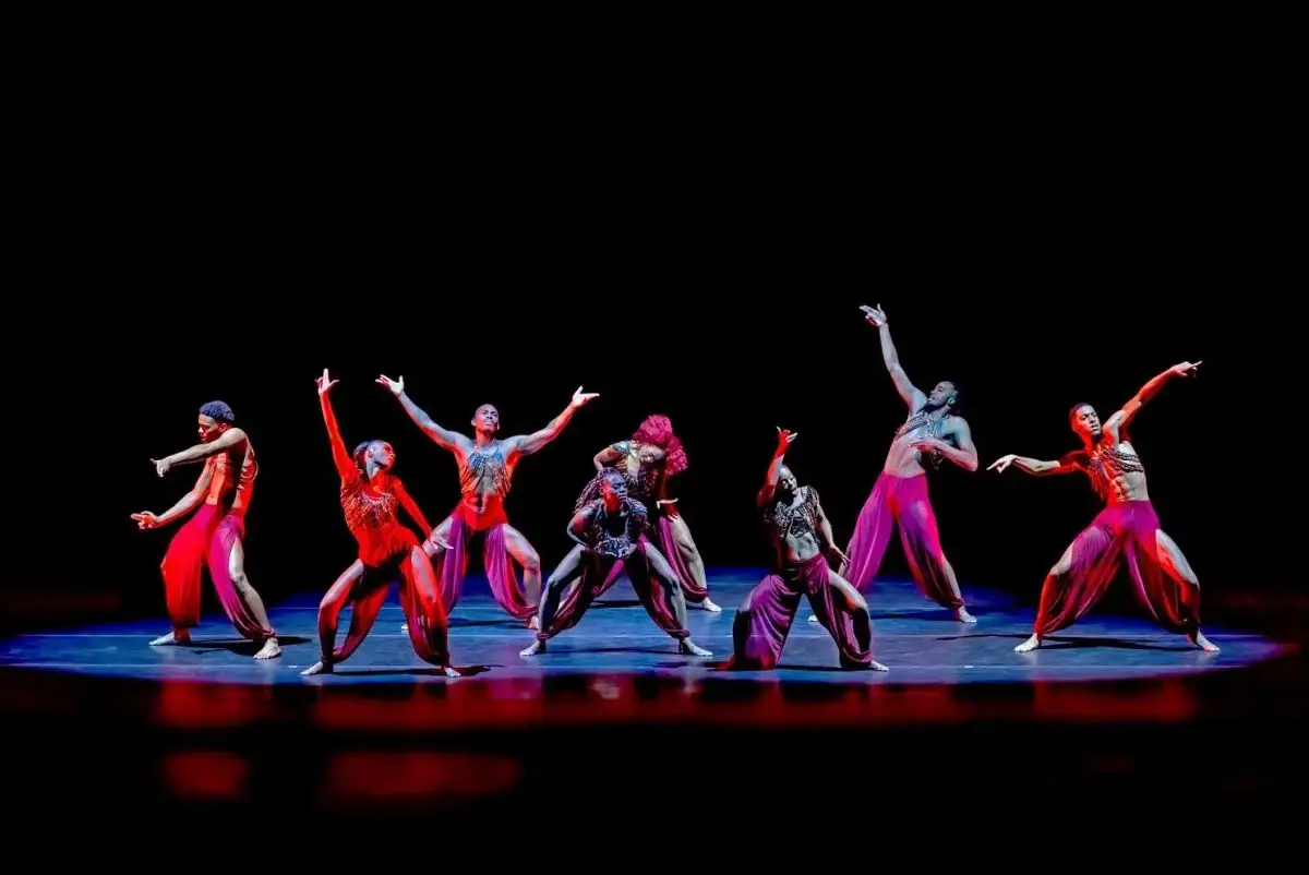 Eight dancers in red and purple costumes perform expressive poses on a dark stage under dramatic lighting.