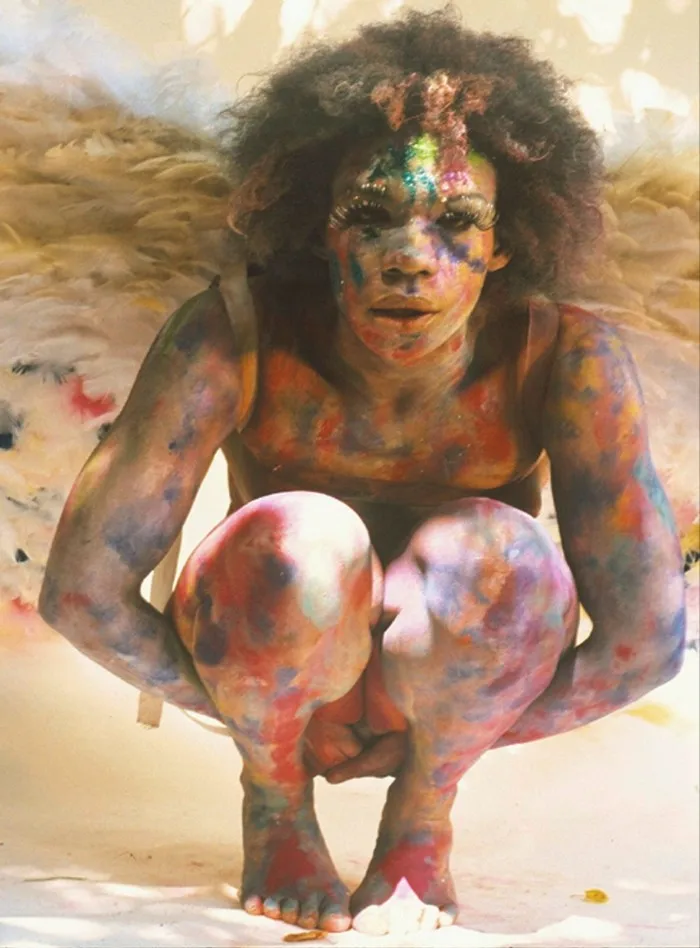 A painting depicts a woman with multicoloured blotches on her skin and elongated eyelashes squatting in what appears to be a hot and dusty environment
