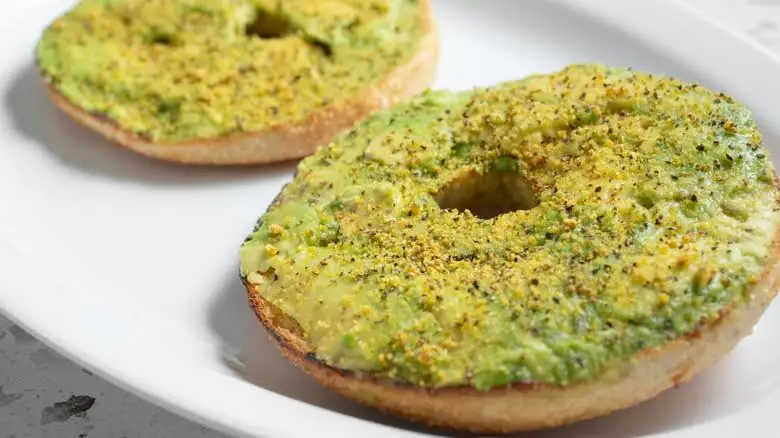Two plated halves of a bagel covered in avocado spread and spices