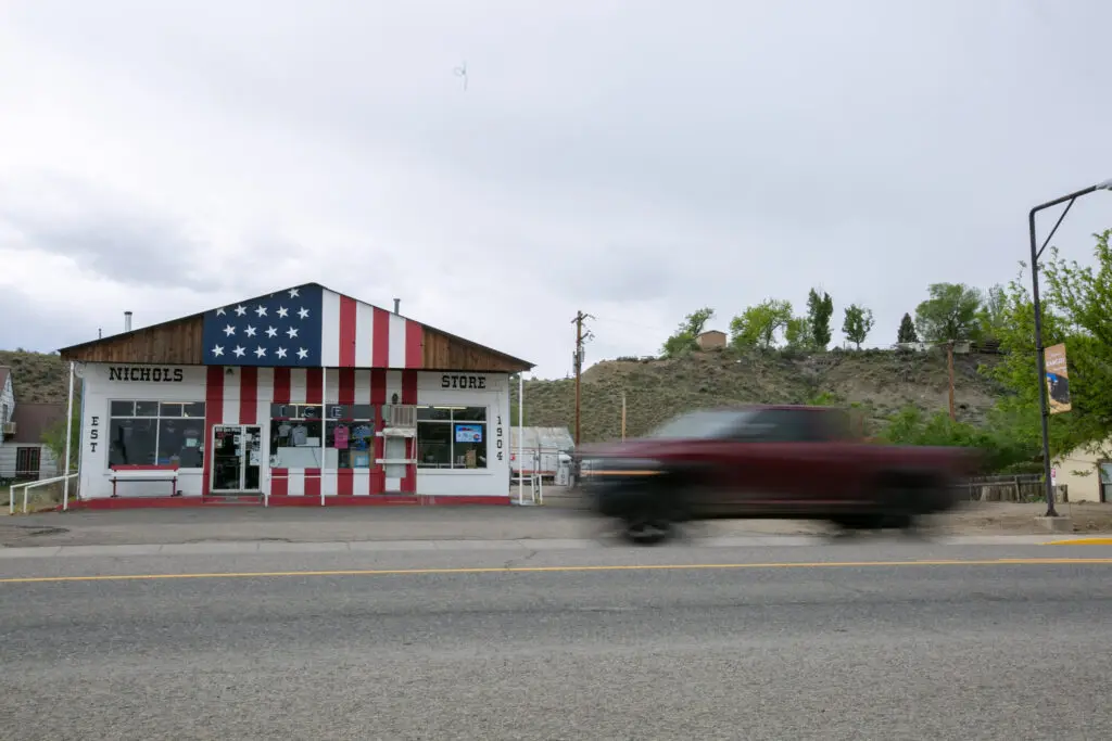 A red pickup truck speeds by a store painted like an American Flag that reads, "NICHOLS STORE."
