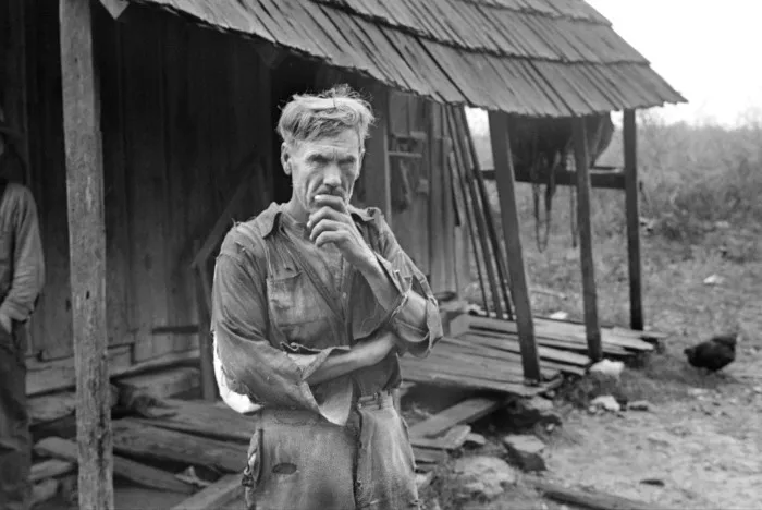 A photograph from the 1930s shows a man in ragged clothing standing with his hand held up to his chin; behind him is a ramshackle house