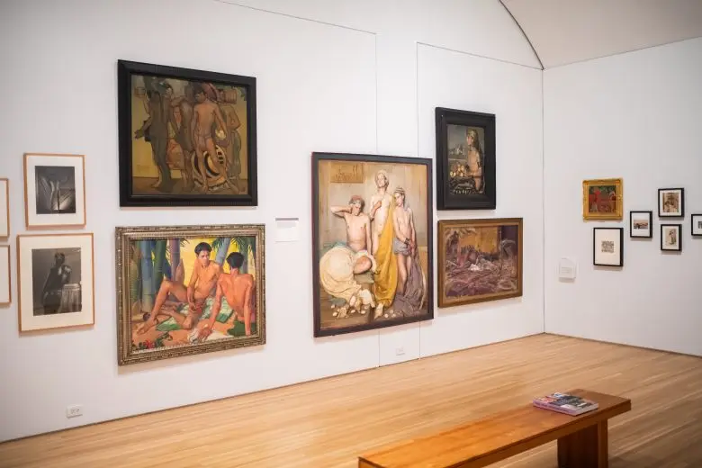 Groups of paintings are hung salon-style on two gallery walls. A cluster of larger works show two or more nude male figures in repose.