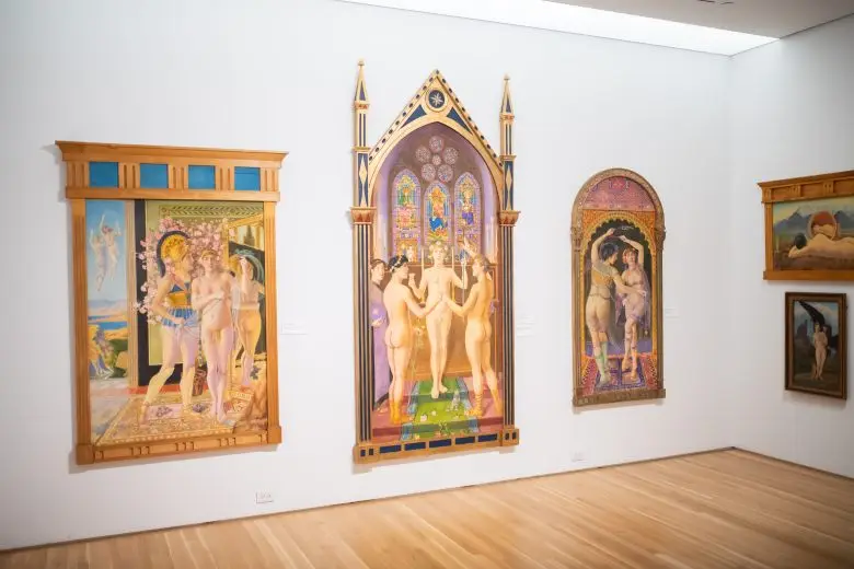 Five large paintings are hung on two white gallery walls, in ornate frames. One is in the shape of a cathedral, one is shaped like an oval doorway. The paintings are very colorful and show various nude figures standing and moving.