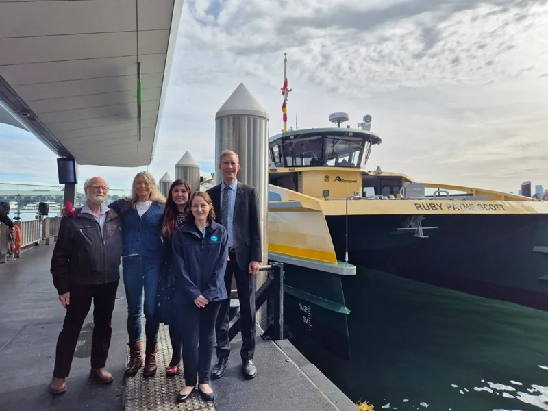 Four people standing in a row on a pier next to a green and yellow Sydney ferry with the name Ruby Payne-Scott painted in white on the ferry.
