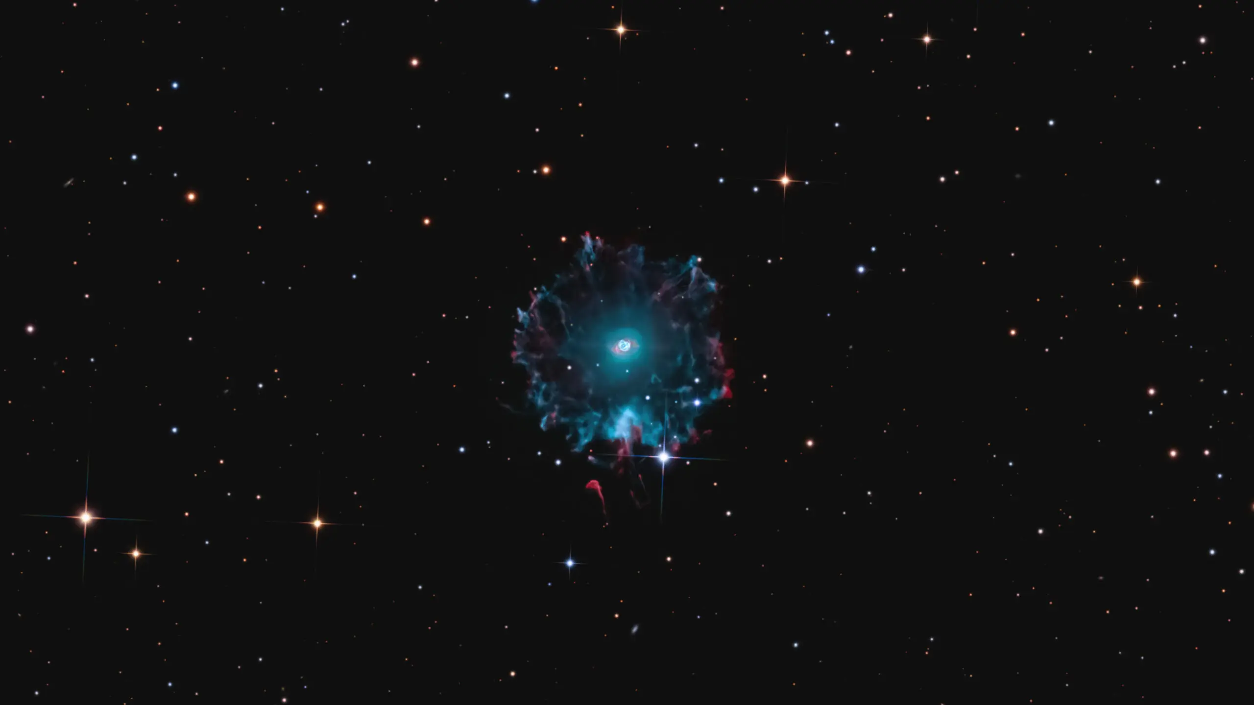 The Cat's Eye Nebula can be seen hanging against a backdrop of stars. The nebula has a wispy blue outer shell and a bright inner core.