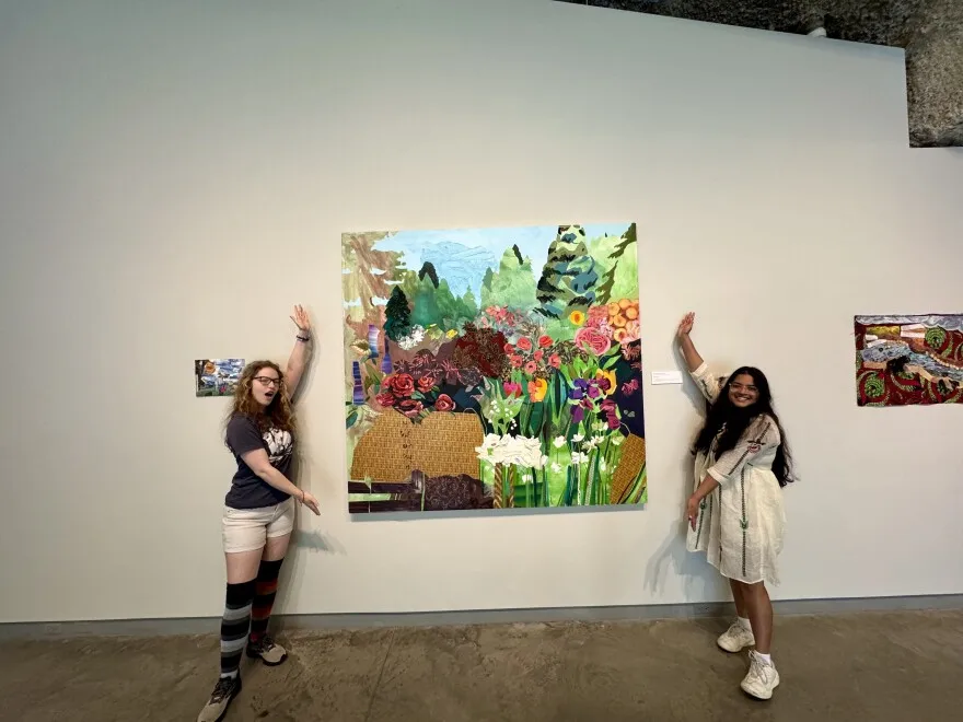 Two women stand on either side of a colorful, nature-themed painting in a gallery, smiling and gesturing toward the artwork on a white wall. One wears shorts and striped socks, the other a white dress.