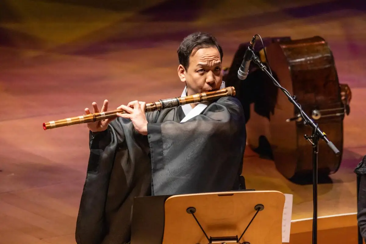 A man in traditional Korean clothing plays a daegeum, a large wooden flute.