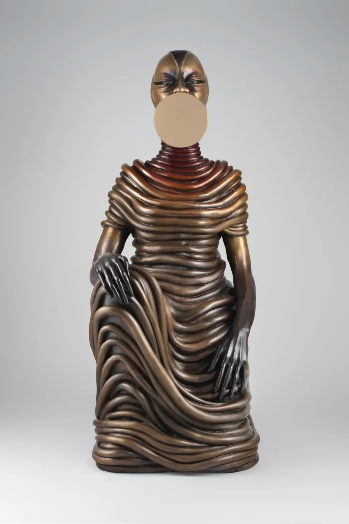 A bronze figure of a woman with a disc on her face. She is wrapped in a rope-like dress
