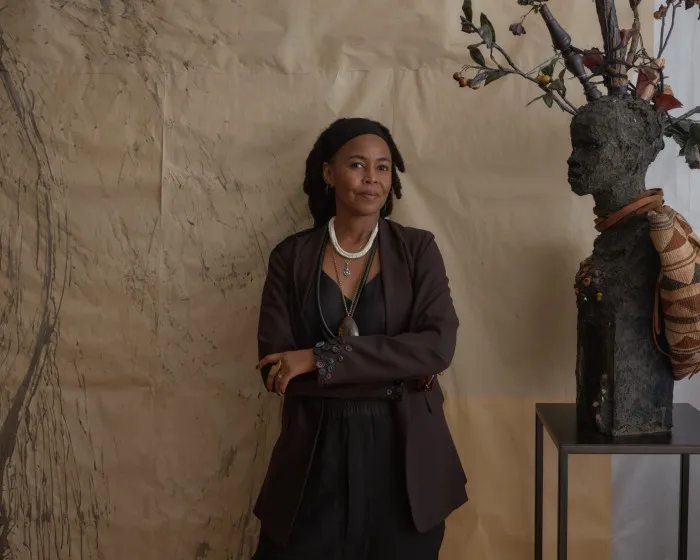 A woman with her arms folded leans against a wall standing near a statue of a bust with twigs and wood coming out of its head. The woman is wearing a silver chain and pendant, as well as a stylish jacket with lots of buttons on the cuffs