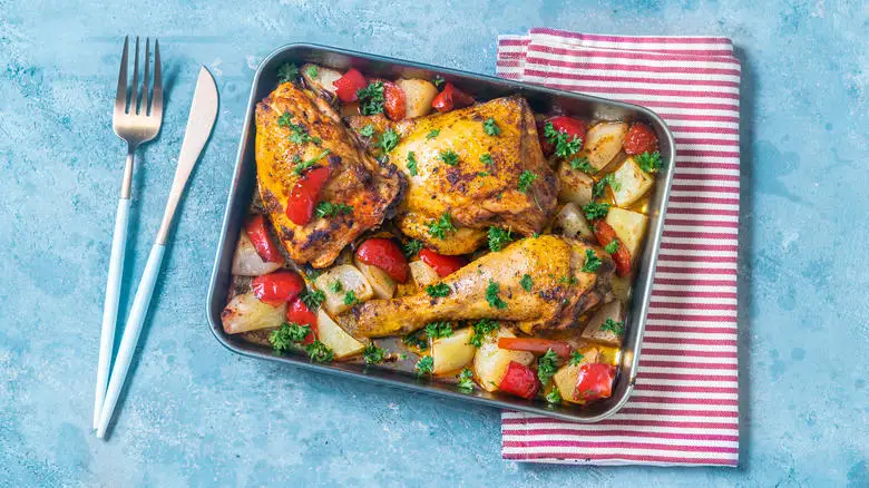 Tray of roast chicken on a bed of root veggies next to a napkin and silverware