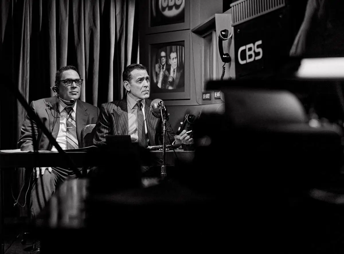 Two men in suits sit behind a desk with microphones. Screens are seen behind them.