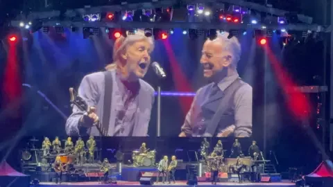 Sebastian Ayling A big screen and stage, showing Sir Paul McCartney signing into a microphone and holding a guitar. Bruce Springsteen is standing to his right holding a guitar and smiling, There is red and blue lighting on stage and there are other musicians.