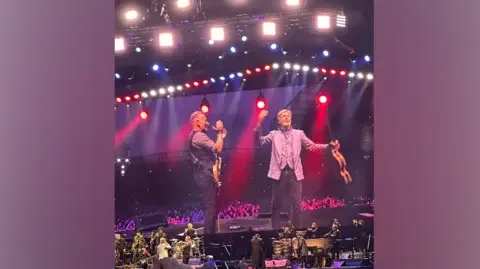 RossEFC95 / X A big screen and stage, showing Sir Paul McCartney holding his hands up to the crowd and holding a guitar. Bruce Springsteen is standing to his left clapping, holding a guitar and smiling. There is red and blue lighting on stage with other musicians. 