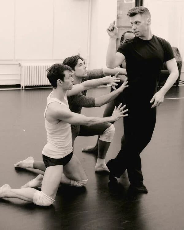 A man, dressed in black casual clothes, stands upright, his body twisting. Two dancers, kneeling on the ground reach out to touch him.