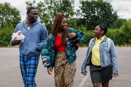 A young black man holding a chicken looks at a blck female friend, with another black female friend in the middle 